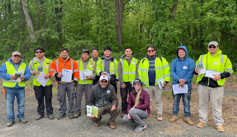 Crews bilingual DC A group of outdoor workers and trainers pose together in front of a wooded area. Most are wearing high-visibility vests and holding informational materials for an educational session related to landscaping. Two facilitators kneel in front