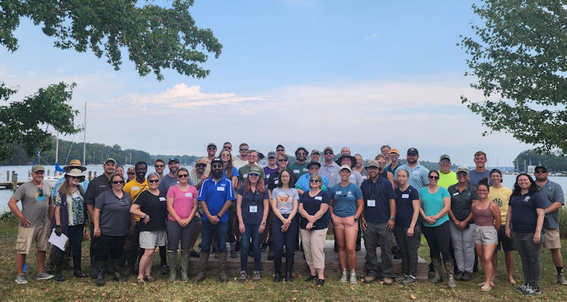 Shorelines MD class A group of adults stand together on a grassy area near a shoreline, participating in a field training session. They are part of the Chesapeake Bay Landscape Professional (CBLP) Shorelines program in Maryland, learning about living shoreline practices. The background features a body of water and natural vegetation, illustrating the coastal environment of the training site